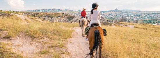 Personen reiten auf Pferden durch eine weite, grüne Landschaft in einer hügeligen Region. Im Hintergrund erstreckt sich eine pittoreske Stadt mit felsigen Formationen unter einem weitläufigen Himmel.
