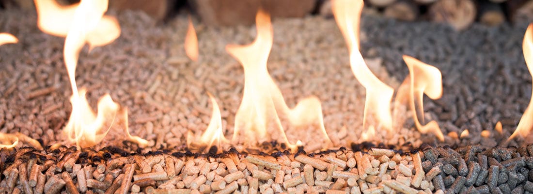 Feuer brennt auf einer Schicht von Holzpellets, die sich auf einem Boden ausbreiten. Der Hintergrund zeigt unscharfe Holzelemente.
