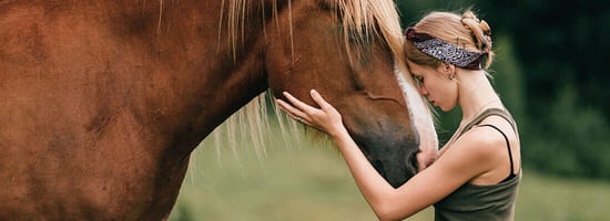 Eine Frau lehnt liebevoll ihren Kopf an den eines Pferdes. Beide befinden sich in einer grünen, natürlichen Umgebung. Das Bild vermittelt Ruhe und Verbundenheit zur Natur.