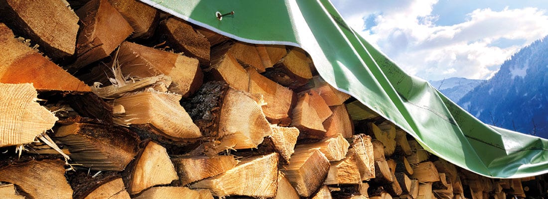 Gestapelte Holzscheite unter einer grünen Plane, perfekt für den Wintervorrat im Gartenbau. Im Hintergrund sind bewaldete Berge und blauer Himmel mit Wolken zu sehen.