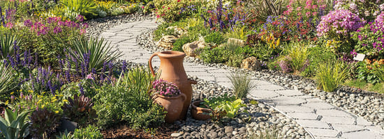 Steinpfad schlängelt sich durch blühenden Garten. Terrakottatöpfe mit bunten Blumen stehen neben dem Weg. Üppige Vegetation und bunte Blüten umgeben den Pfad, der durch den sonnigen Garten führt.