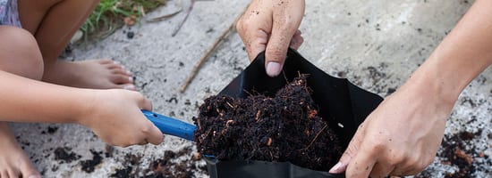 Erwachsene und Kind befüllen gemeinsam einen Pflanzsack mit Erde, umgeben von Gartenboden. Die Szene zeigt gemeinsames Gärtnern im Freien, ideal für Gartenbauaktivitäten und nachhaltige Erdbeschaffung.