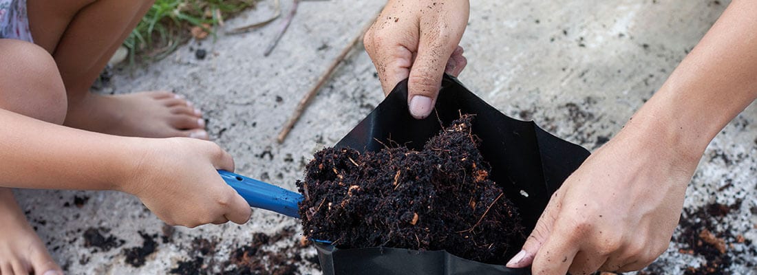 Erwachsene und Kind befüllen gemeinsam einen Pflanzsack mit Erde, umgeben von Gartenboden. Die Szene zeigt gemeinsames Gärtnern im Freien, ideal für Gartenbauaktivitäten und nachhaltige Erdbeschaffung.
