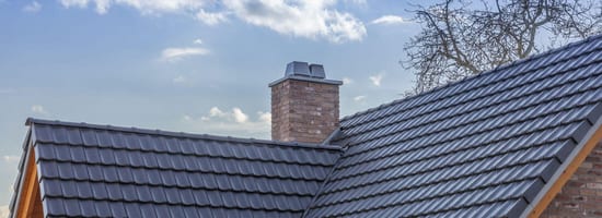 Dach mit grauen Ziegeln; Schornstein aus Backstein; blauer Himmel im Hintergrund; kahler Baum rechts.