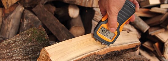 Hand hält Feuchtigkeitsmesser auf Holzscheit; Anzeige zeigt 20,9%. Hintergrund: gestapeltes Brennholz. Ideale Vorbereitung von Holzbriketts für effiziente Wärmeproduktion im Heizbereich.