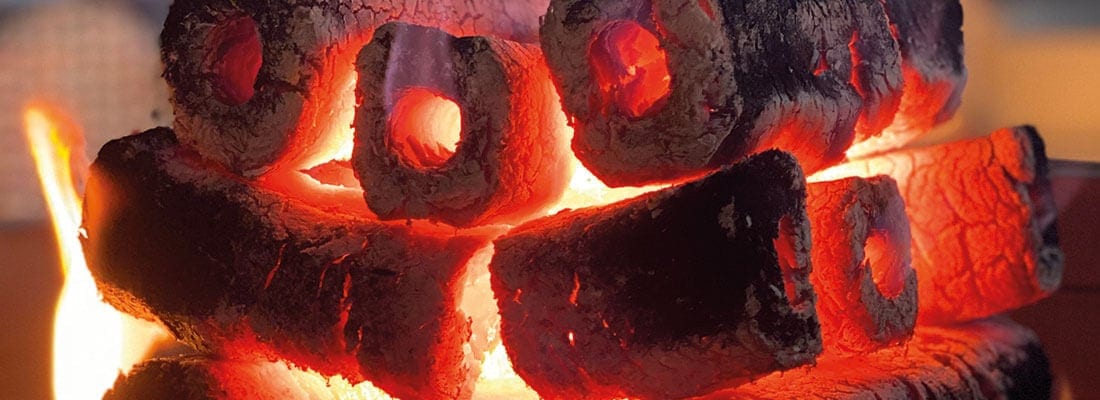 Glühende Holzbriketts mit zentralen Löchern liegen übereinander und brennen intensiv. Der Hintergrund ist unscharf und sorgt für eine warme, einladende Atmosphäre.
