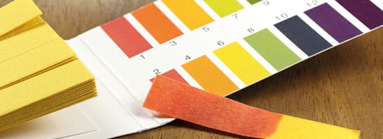 pH-Teststreifen-Set, das verschiedene Farbstufen von 1 bis 14 zeigt, liegt auf einem Holztisch. Ein einzelner Streifen zeigt eine orangefarbene Verfärbung, die auf den Säuregrad hindeutet.