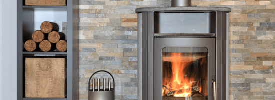 Moderner Kaminofen mit sichtbarer Flamme in gemütlicher Ziegelstein-Umgebung, daneben gestapelte Holzbriketts. Perfekt für nachhaltige Wärme in Wohnräumen.