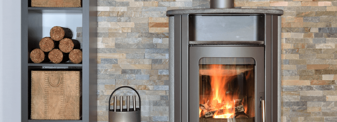 Moderner Kaminofen mit sichtbarer Flamme in gemütlicher Ziegelstein-Umgebung, daneben gestapelte Holzbriketts. Perfekt für nachhaltige Wärme in Wohnräumen.