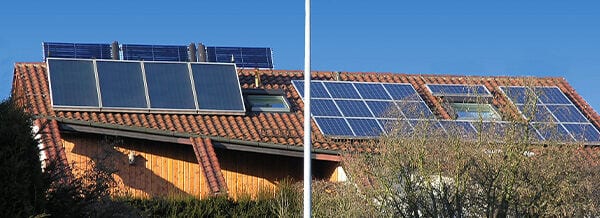 Solaranlagen auf einem Ziegeldach eines Hauses, die Sonnenenergie in Strom umwandeln. Umgebung ist blauem Himmel und Bäumen geprägt. Fokus liegt auf erneuerbarer Energie durch Solarpanels.